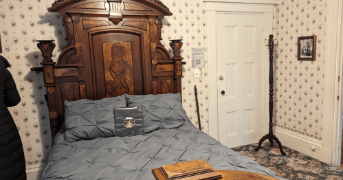 A vintage bedroom featuring a wooden bed, blue bedding, floral wallpaper, and a door with a coat rack nearby.