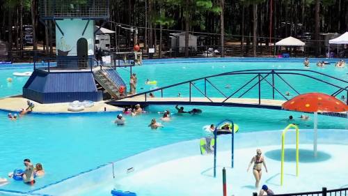 A busy outdoor pool with people swimming, a slide, and a colorful mushroom-shaped structure. Trees in the background.
