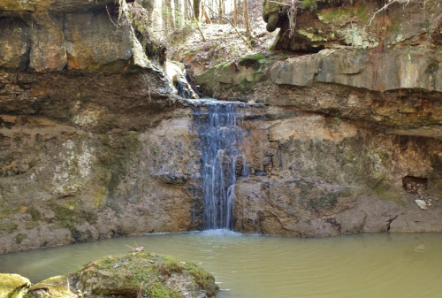 A small waterfall cascades over rocky cliffs into a tranquil pool surrounded by lush greenery.