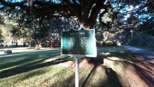 A historical marker for Logtown stands under a large tree, surrounded by greenery and a dirt road.