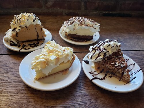 Four slices of pie on white plates, topped with whipped cream, chocolate drizzle, and nuts, on a wooden table.