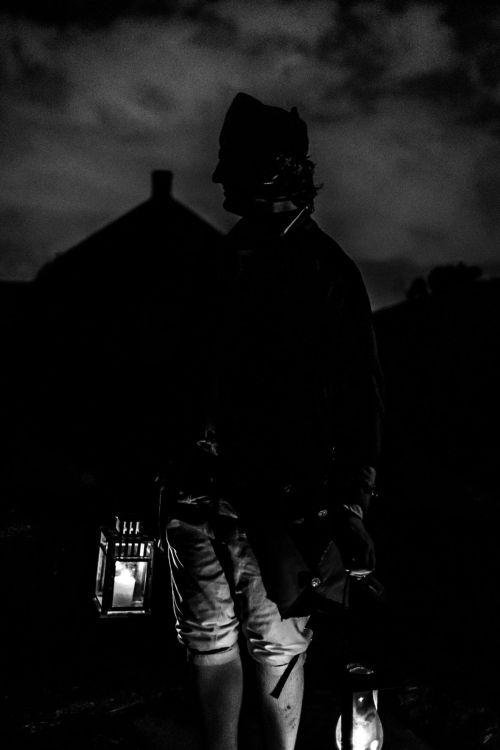 A silhouetted figure holding a lantern stands against a dark, cloudy sky, with a building faintly visible in the background.