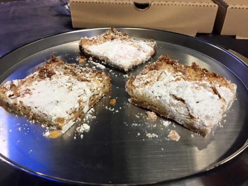 Three pieces of dessert bars dusted with powdered sugar on a silver platter, with pizza boxes in the background.
