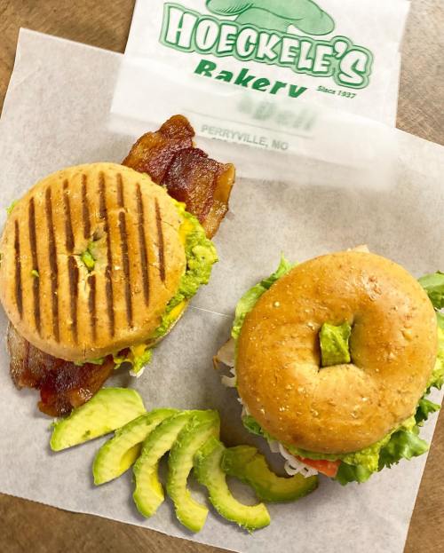Two sandwiches on a paper tray: one with bacon and avocado, the other with lettuce and tomato, alongside sliced avocado.