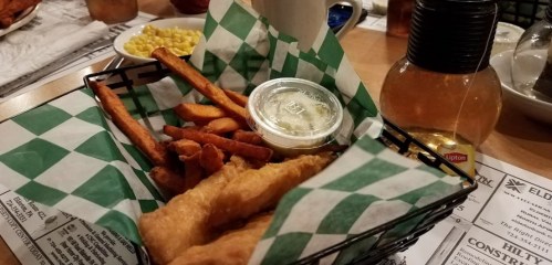 A basket of fish and fries with a side of dipping sauce, served on a checkered paper, with corn in the background.