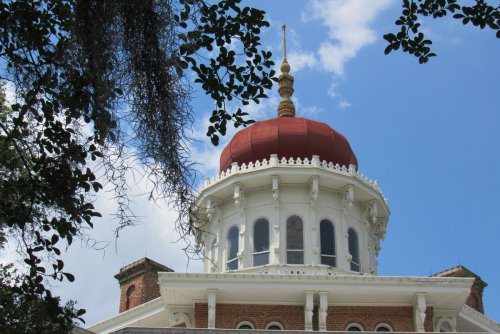 The ornate dome of a historic building, featuring a red roof and decorative details, framed by tree branches.