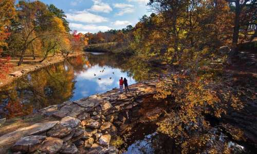 A couple stands on a rocky ledge by a serene river, surrounded by vibrant autumn foliage and ducks swimming nearby.