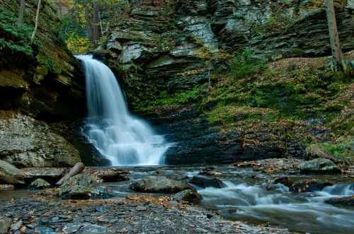A serene waterfall cascades over rocky cliffs, surrounded by lush greenery and autumn leaves.