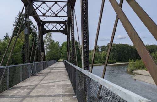 A metal bridge spans a river, surrounded by trees and greenery under a partly cloudy sky.