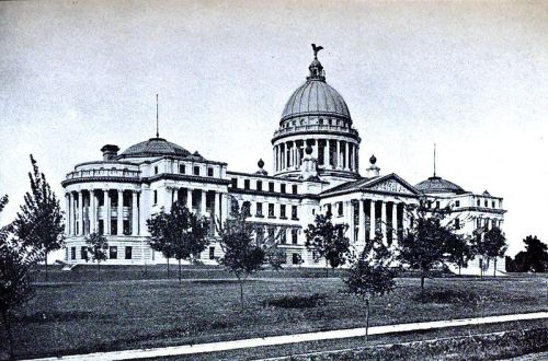 Historic building with a large dome, columns, and surrounding trees, set in a landscaped area.