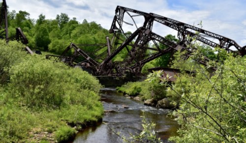 Rusty, abandoned metal structures lean over a serene stream, surrounded by lush green trees and a clear blue sky.