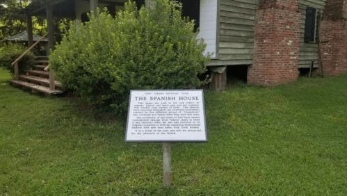 A historical sign titled "The Spanish House" in front of a rustic building surrounded by greenery.