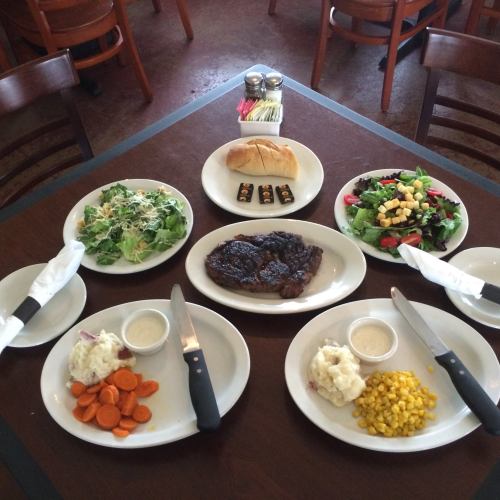 A table set with a steak, sides of mashed potatoes, carrots, corn, salads, and a bread roll.
