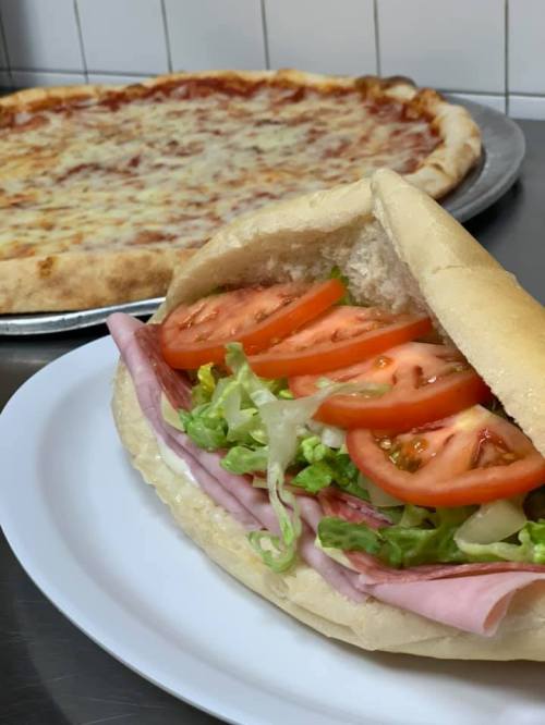 A close-up of a sandwich with ham, lettuce, and tomato on a plate, with a pizza in the background.