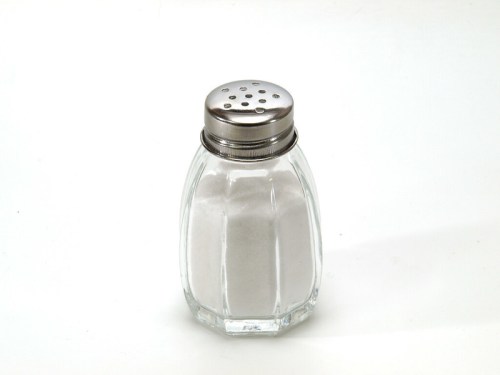 A clear glass salt shaker with a silver lid, filled with white salt, on a white background.