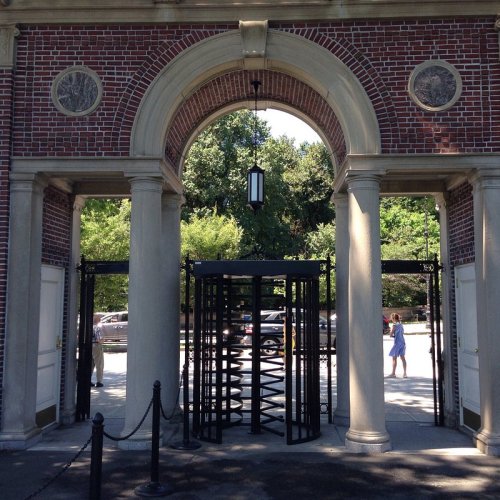 A grand entrance with a large archway, flanked by decorative pillars and a revolving door, surrounded by greenery.