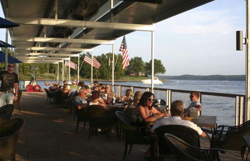 Outdoor dining area by a lake, with people enjoying meals and drinks, American flags, and a boat in the background.
