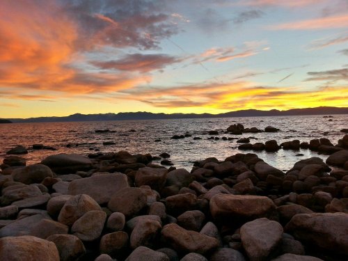 A serene sunset over a rocky shoreline, with vibrant orange and purple clouds reflecting on the water.