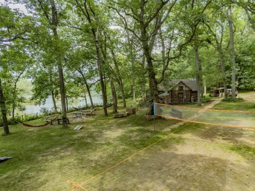 A serene outdoor scene featuring a cabin by a river, surrounded by trees and a grassy area with hammocks.