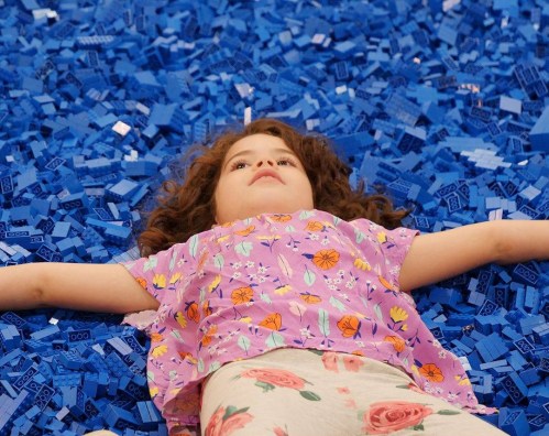 A child lies on a bed of blue LEGO bricks, arms outstretched, with a joyful expression.