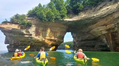 Three kayakers paddle through calm waters near a large rock formation with an arch, surrounded by lush greenery.
