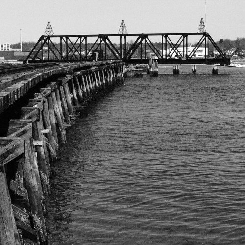 Black and white image of a wooden pier leading to a metal bridge over calm water.