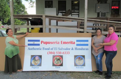 Three women stand beside a sign for "Pupuseria Emerita," promoting authentic Salvadoran and Honduran food.