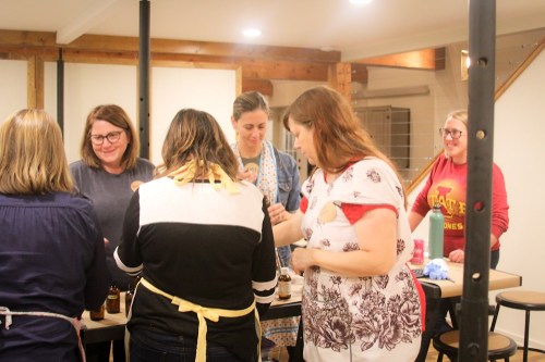 A group of women gather around a table, engaged in a crafting activity, with jars and supplies in a cozy indoor setting.