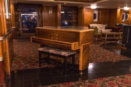 A polished wooden piano with a decorative bench, set in an elegant room with patterned carpet and vintage furnishings.