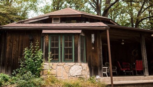 A rustic wooden cabin with a stone foundation, surrounded by trees and greenery.