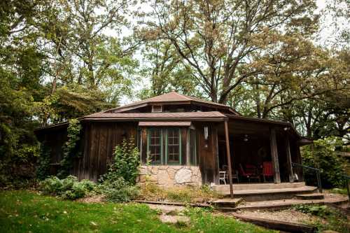 A rustic wooden cabin surrounded by trees, featuring a porch and large windows, set in a lush green landscape.