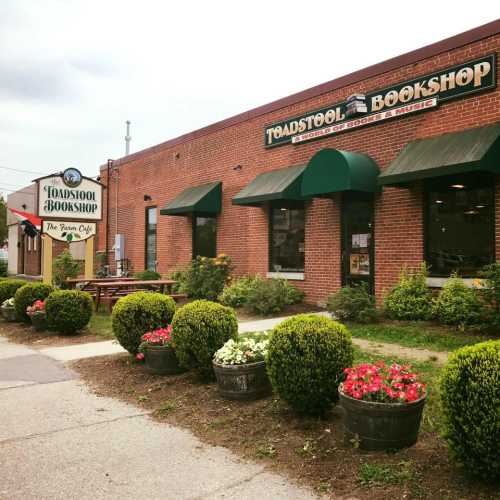 A brick building with green awnings houses Toadstool Bookshop, surrounded by colorful flower beds and shrubs.