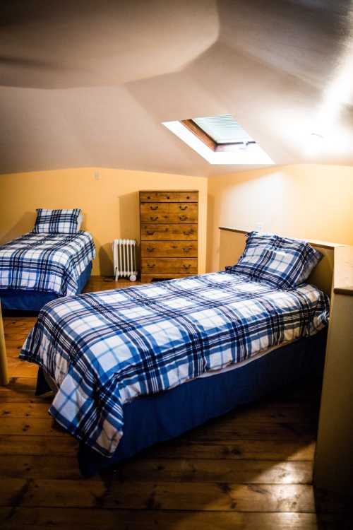 Cozy attic bedroom with two twin beds, plaid bedding, a wooden dresser, and a skylight. Warm, inviting atmosphere.