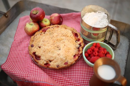 A freshly baked pie on a red checkered cloth, surrounded by apples, flour, raspberries, and a cup of milk.