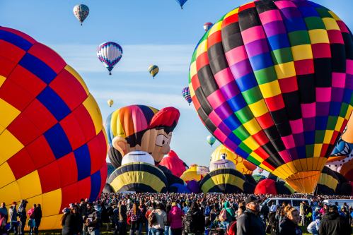 Colorful hot air balloons fill the sky above a large crowd at a festival, with cartoon characters among the balloons.
