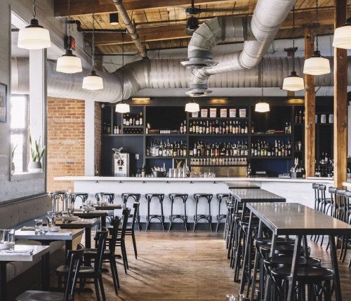 Modern restaurant interior with wooden beams, a bar, and black chairs arranged around tables. Warm, inviting atmosphere.