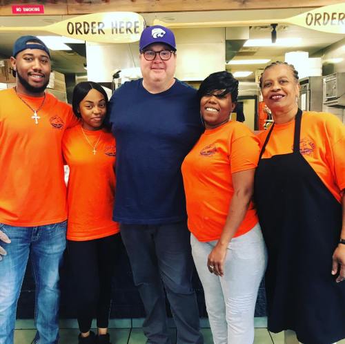 A group of five people, wearing orange shirts, smile together in a restaurant setting.