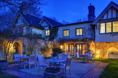 A charming house at dusk, featuring warm lights, a patio with tables, and lush greenery surrounding the outdoor area.
