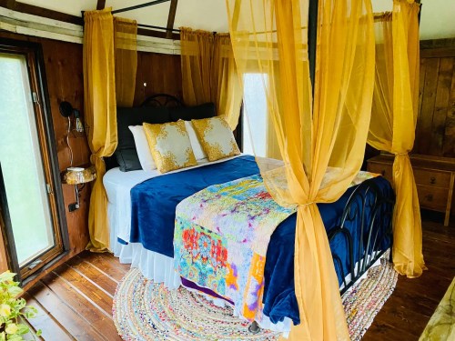 Cozy bedroom with a canopy bed, colorful blankets, and warm yellow drapes, surrounded by wooden walls and large windows.