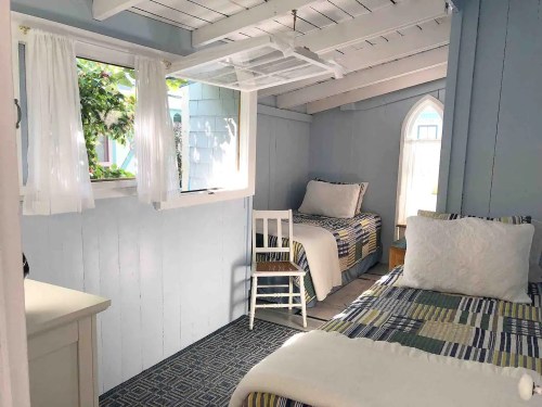 Cozy bedroom with two beds, a chair, and bright windows, featuring light blue walls and natural light.