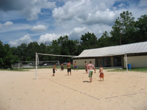 A group of people playing beach volleyball on a sandy court with a building and trees in the background.
