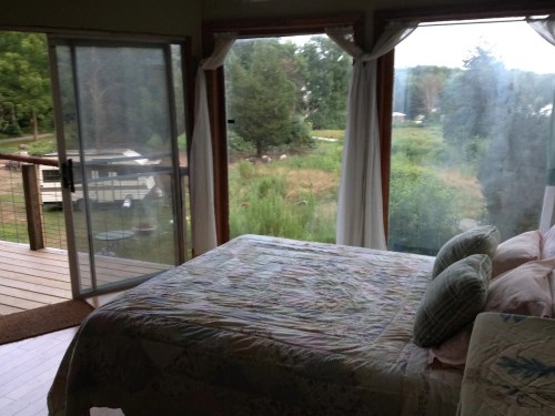 Cozy bedroom with a quilted bedspread, large windows overlooking a green landscape, and a sliding glass door.