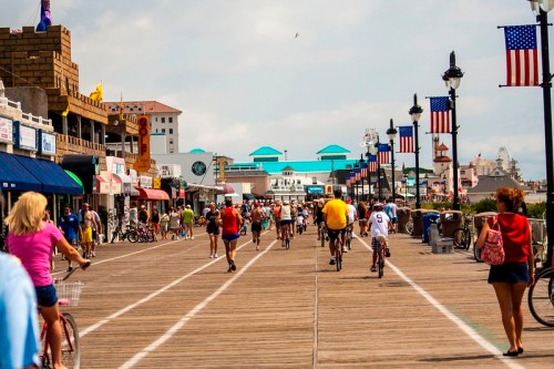 A busy boardwalk scene with people biking and walking, shops lining the path, and flags waving in the breeze.