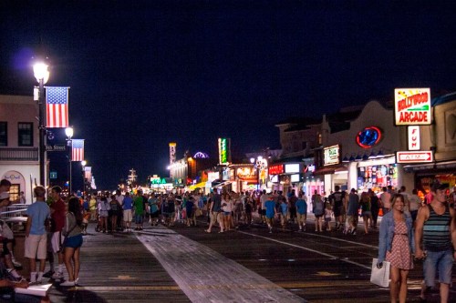 A bustling boardwalk at night, filled with people and colorful neon signs from shops and arcades.