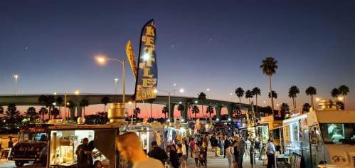 A bustling food truck event at dusk, with palm trees and a colorful sky in the background.