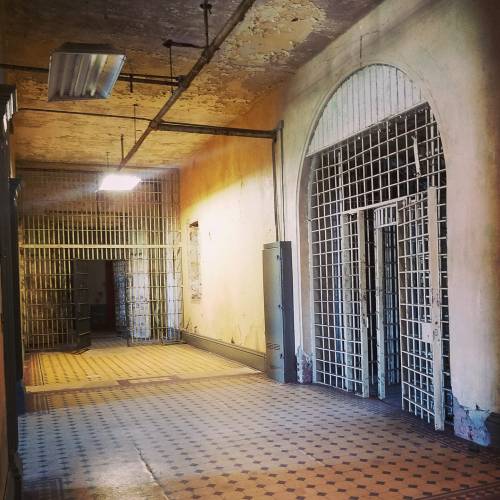 A dimly lit corridor in an old prison, featuring barred doors and peeling walls.