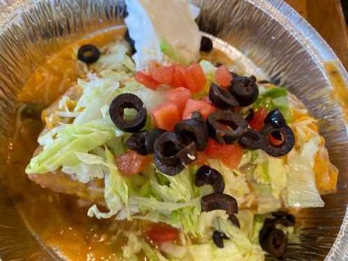 A foil container filled with a burrito topped with lettuce, tomatoes, black olives, and cheese in a savory sauce.