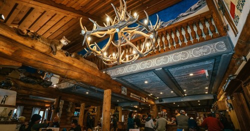 A rustic restaurant interior featuring wooden beams, an antler chandelier, and patrons enjoying their meals.