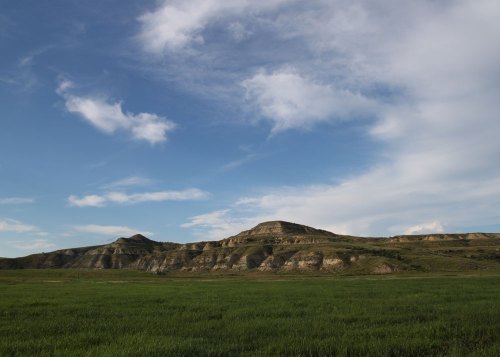 A wide view of rolling hills under a blue sky with scattered clouds, surrounded by green grassland.