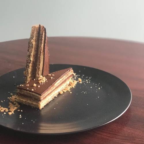 A slice of layered chocolate cake on a black plate, garnished with crumbs, set on a wooden table.